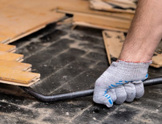A person wearing gloves is using a metal pry bar to lift old wood flooring off a dusty black subfloor during renovation.