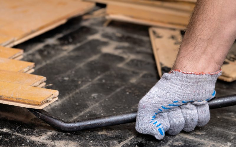 A person wearing gloves is using a metal pry bar to lift old wood flooring off a dusty black subfloor during renovation.