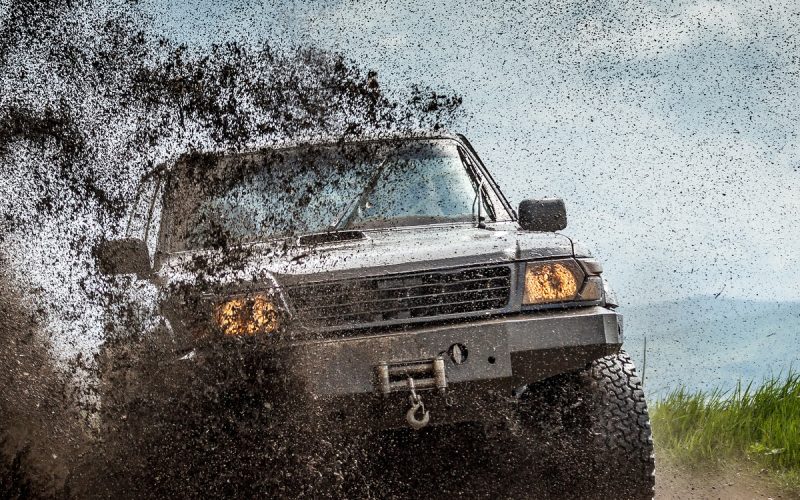 A large truck driving fast on an off-road trail during the daytime. Soil flies off the terrain as the truck drives.