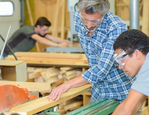 A man in safety glasses guiding a wood board through a table saw while another person watches in a wood shop.