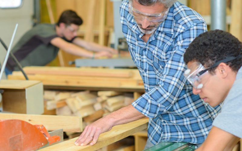 A man in safety glasses guiding a wood board through a table saw while another person watches in a wood shop.