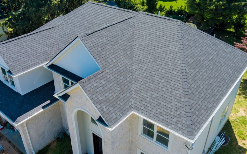 A large house with gray asphalt shingle roofs, stone siding, white trim, and a large yard near the property.