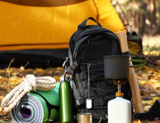 A yellow tent stands behind a black backpack and camping gear arranged on fallen leaves in a wooded campsite.