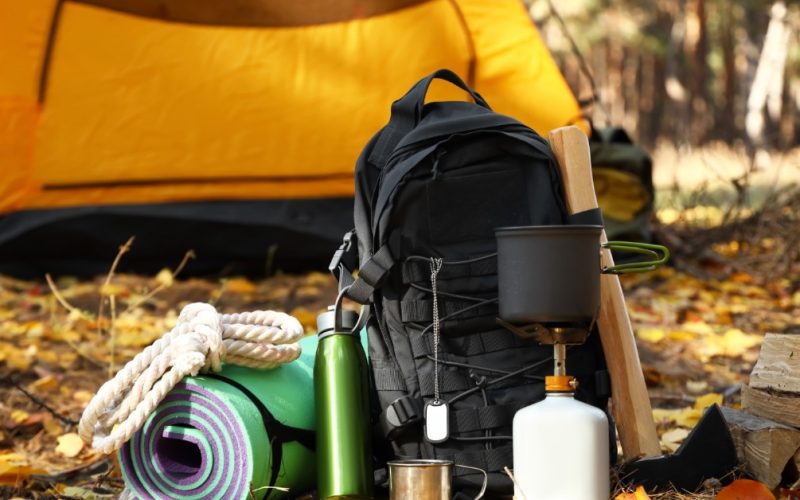 A yellow tent stands behind a black backpack and camping gear arranged on fallen leaves in a wooded campsite.
