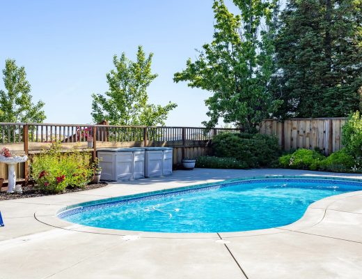 A curved backyard swimming pool surrounded by a concrete patio, green lawn, fence, trees, and blue chairs.