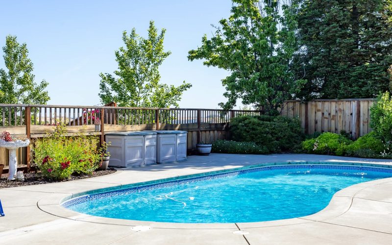 A curved backyard swimming pool surrounded by a concrete patio, green lawn, fence, trees, and blue chairs.
