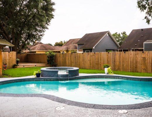 A backyard space with a curved swimming pool, a raised spa, a waterfall feature, green lawn, and a wooden privacy fence.