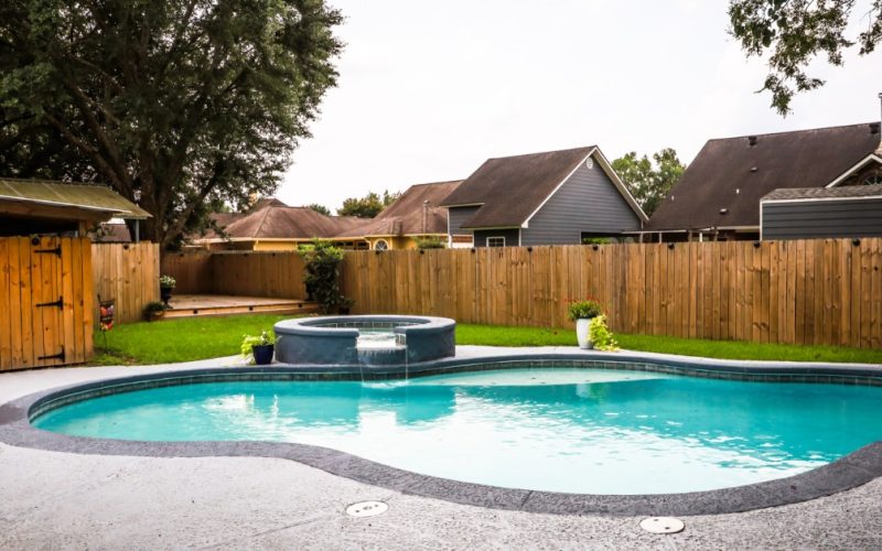 A backyard space with a curved swimming pool, a raised spa, a waterfall feature, green lawn, and a wooden privacy fence.