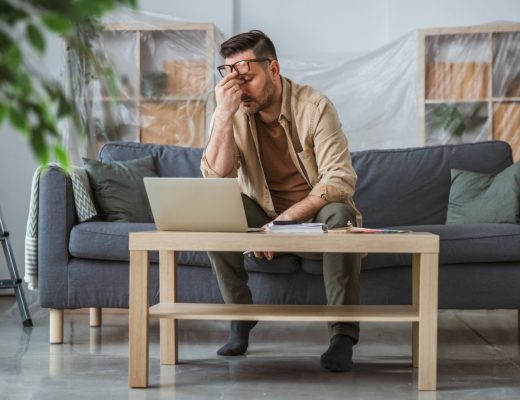 A man sitting on a gray sofa, holding his glasses and frustrated about home renovations, with covered shelves nearby.