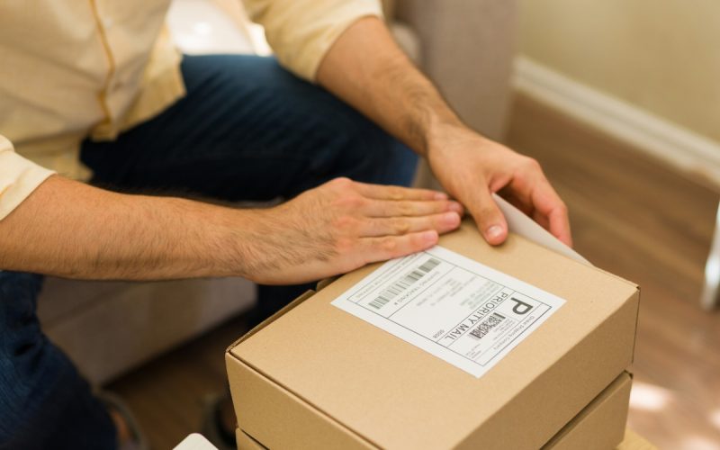 An home-based entrepreneur pressing a shipping label onto a cardboard box beside papers on a wooden table.