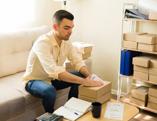 A man attaching a shipping label to a cardboard box while sitting beside shelves filled with packaged boxes.