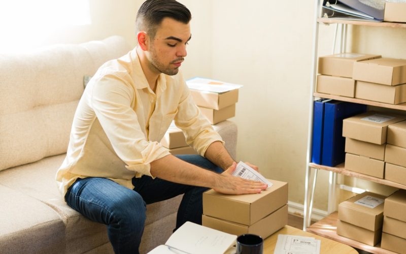 A man attaching a shipping label to a cardboard box while sitting beside shelves filled with packaged boxes.