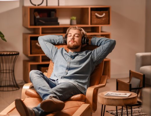 A close-up of a man wearing headphones, sitting in an armchair with his eyes closed in a bright living room.