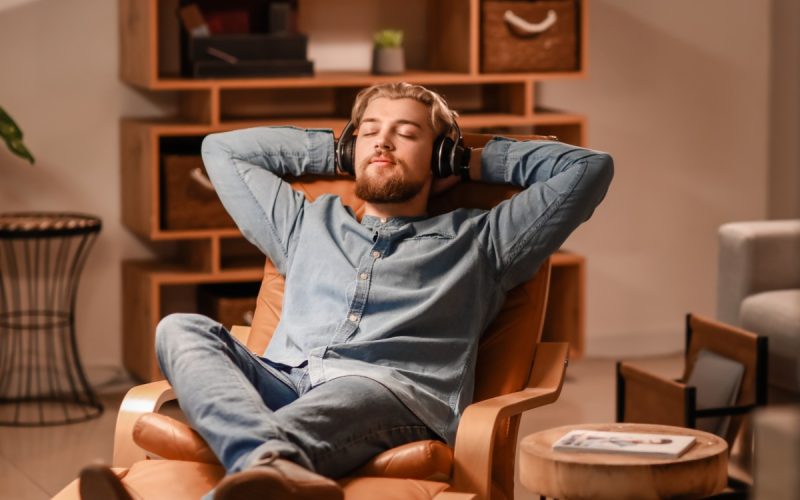 A close-up of a man wearing headphones, sitting in an armchair with his eyes closed in a bright living room.