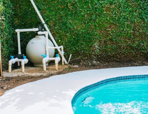 A swimming pool next to an equipment pad with a sand filter, white pipes, and valves installed beside a dense green hedge.