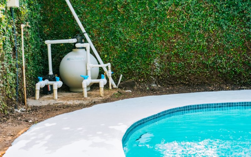 A swimming pool next to an equipment pad with a sand filter, white pipes, and valves installed beside a dense green hedge.