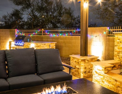 An outdoor patio at night with cushioned seating, a fire table, string lights, a grill, and an illuminated waterfall.