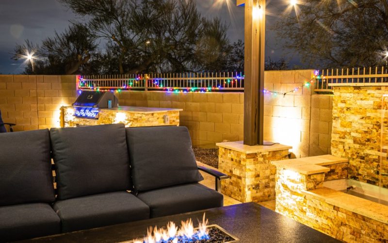 An outdoor patio at night with cushioned seating, a fire table, string lights, a grill, and an illuminated waterfall.