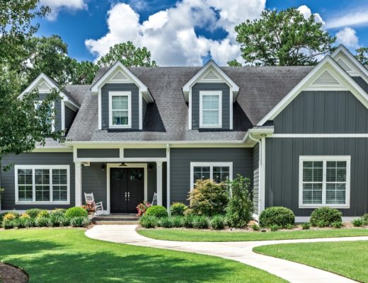 A large gray house with white trim, dormer windows, a neatly landscaped lawn, and a curved front walkway.