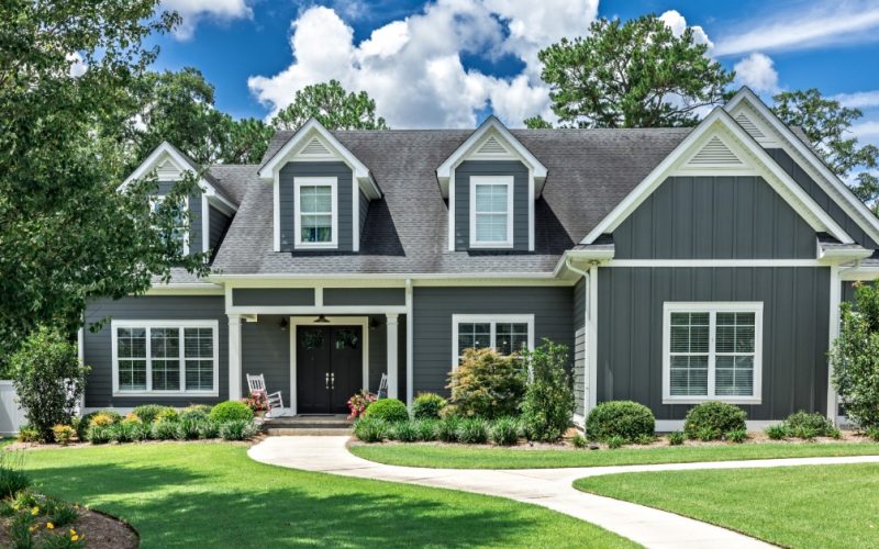 A large gray house with white trim, dormer windows, a neatly landscaped lawn, and a curved front walkway.