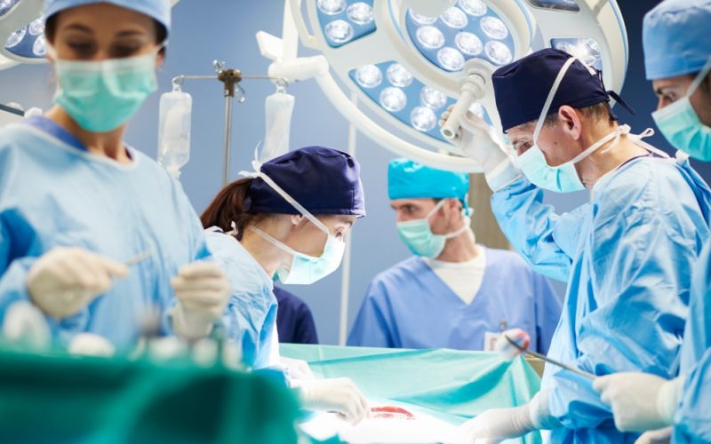 A surgical team wearing masks, scrubs, and gloves while performing an operation under bright surgical lights.