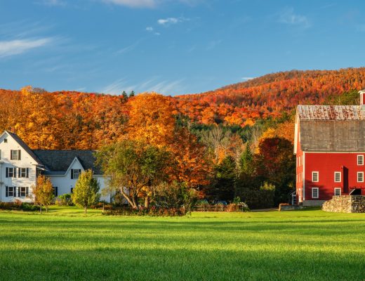 A white farmhouse and a large red barn beside a green field with colorful autumn trees and hills in the background.