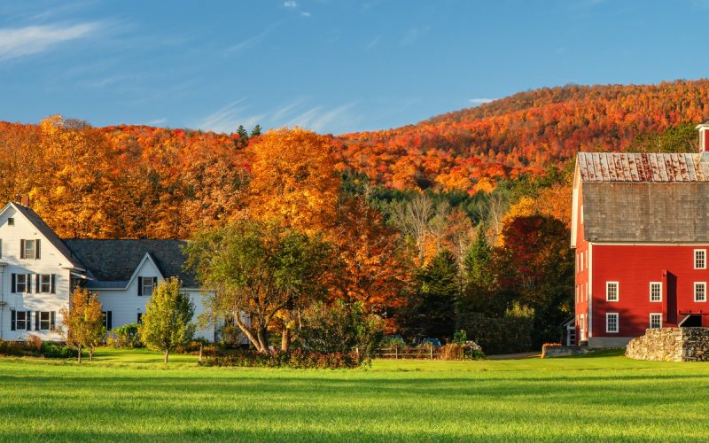A white farmhouse and a large red barn beside a green field with colorful autumn trees and hills in the background.