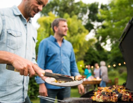 A man placing grilled skewers on a plate near another man and a barbecue with people at a garden table behind.
