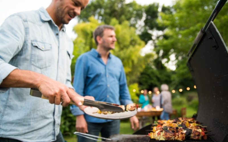A man placing grilled skewers on a plate near another man and a barbecue with people at a garden table behind.