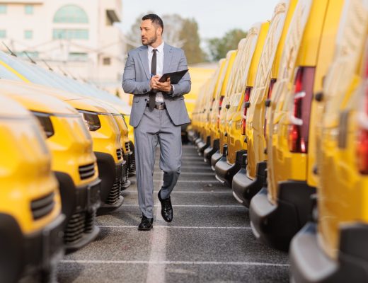 A man in a gray suit holding a clipboard while walking between rows of yellow delivery vans in a parking lot.