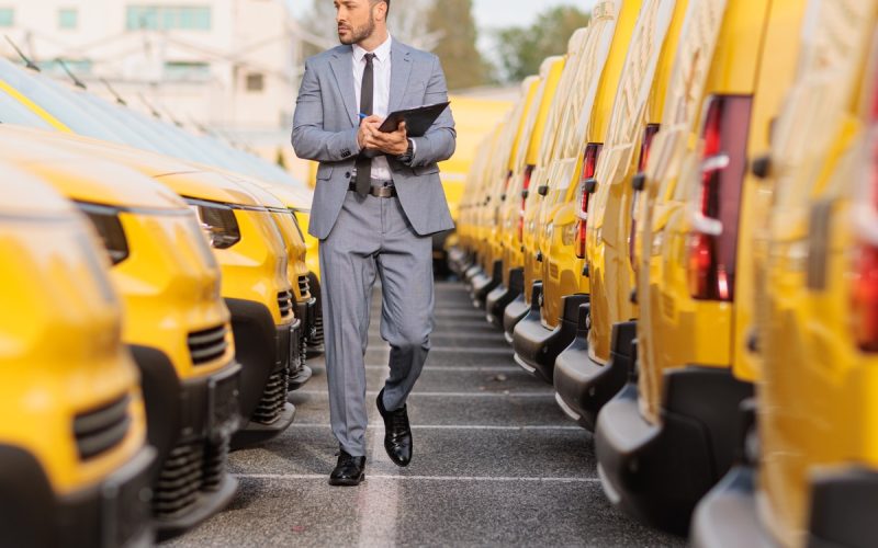 A man in a gray suit holding a clipboard while walking between rows of yellow delivery vans in a parking lot.