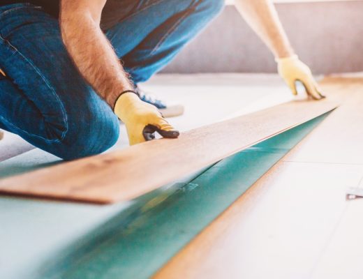 A man is laying laminate flooring that is in the style of wood boards. The man is wearing gloves as he lays the boards.