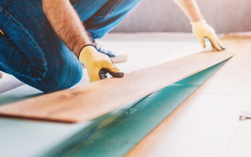 A man is laying laminate flooring that is in the style of wood boards. The man is wearing gloves as he lays the boards.