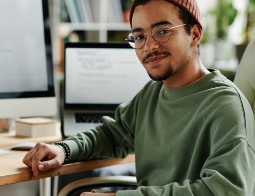 Man wearing glasses and a knit cap sits at a desk with a desktop monitor and laptop in a bright home office.