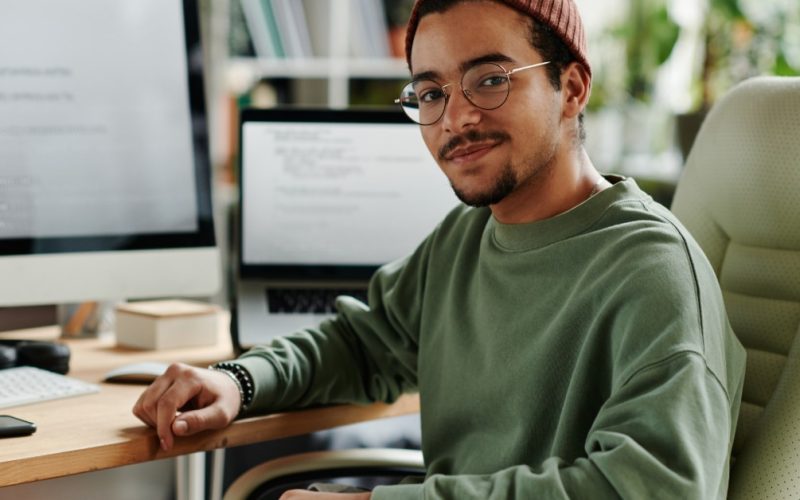 Man wearing glasses and a knit cap sits at a desk with a desktop monitor and laptop in a bright home office.