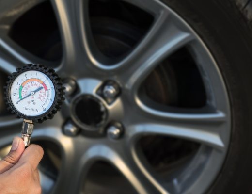 A close-up of a hand holding a tire pressure gauge with the needle at zero in front of a car wheel and tire.