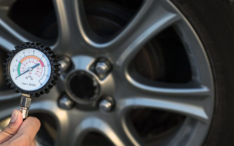 A close-up of a hand holding a tire pressure gauge with the needle at zero in front of a car wheel and tire.