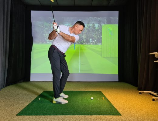 A side view of a man in a white polo and black slacks mid-swing with a golf club inside an indoor golf simulator.