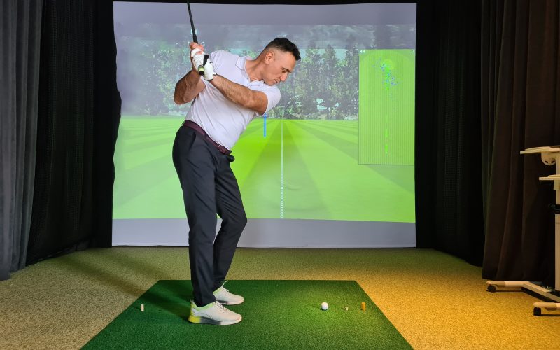 A side view of a man in a white polo and black slacks mid-swing with a golf club inside an indoor golf simulator.