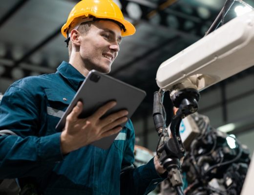 A close-up of a person using a tablet beside robotic arms and industrial machinery inside a manufacturing facility.