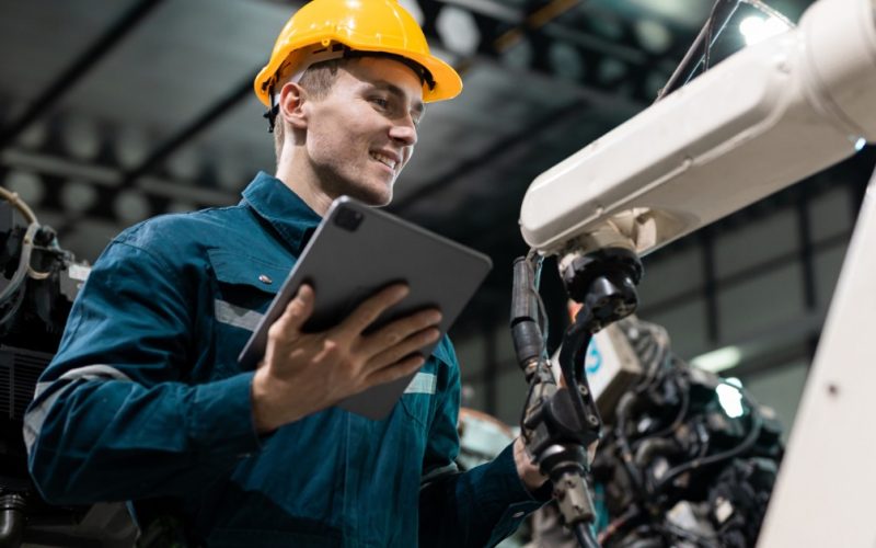 A close-up of a person using a tablet beside robotic arms and industrial machinery inside a manufacturing facility.