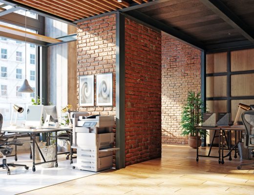 Sunlit office interior with brick partitions, wood ceiling beams, workstations, computers, and rolling desk chairs.