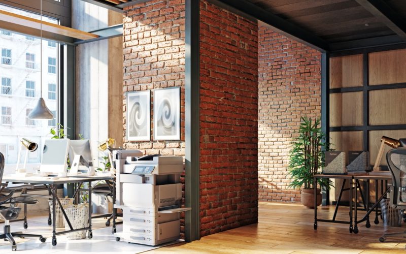 Sunlit office interior with brick partitions, wood ceiling beams, workstations, computers, and rolling desk chairs.