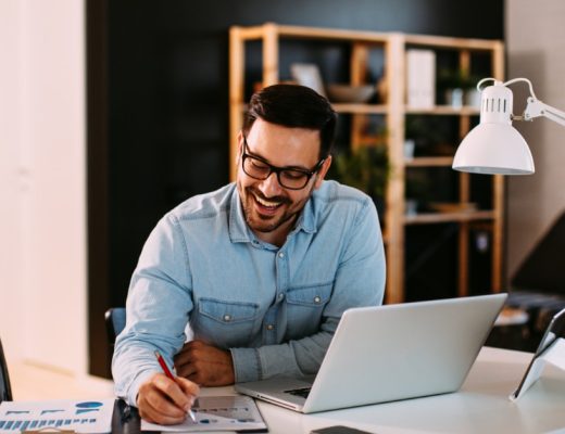  Man wearing glasses reviews business charts at his desk beside an open laptop, smartphone, tablet, and lamp.