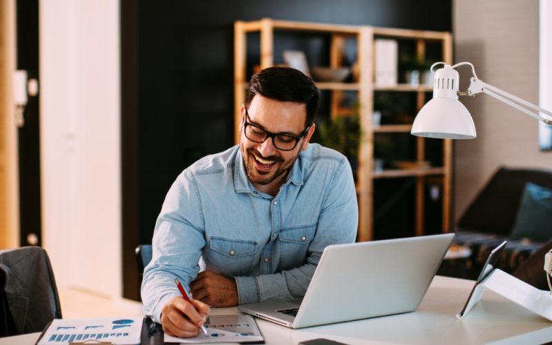  Man wearing glasses reviews business charts at his desk beside an open laptop, smartphone, tablet, and lamp.