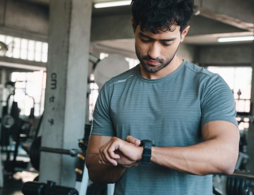 A man is standing in the middle of a fitness center. He’s looking at a watch on his wrist.