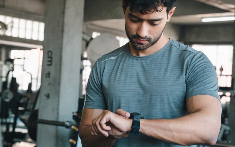 A man is standing in the middle of a fitness center. He’s looking at a watch on his wrist.