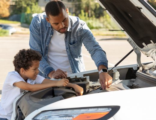 A man and boy standing beside an open car hood, looking at the engine with trees and bright light behind them.