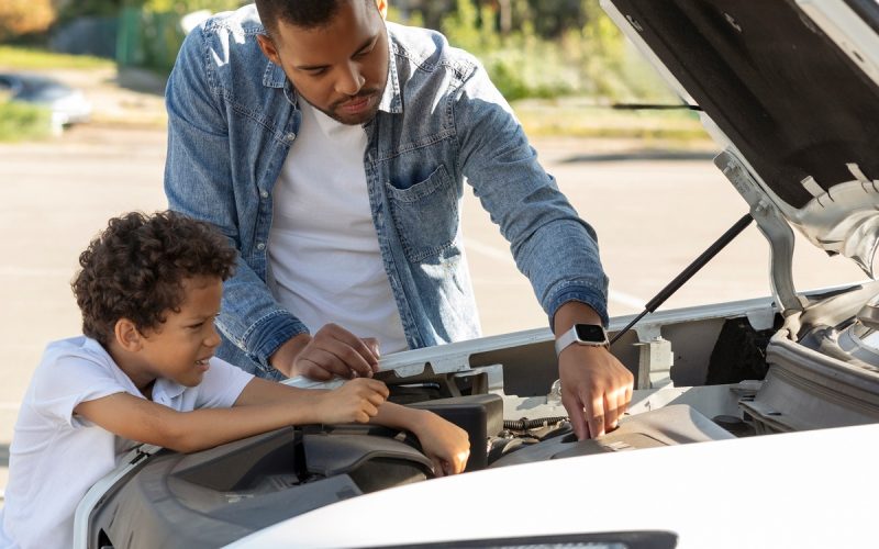 A man and boy standing beside an open car hood, looking at the engine with trees and bright light behind them.