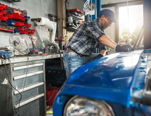 A person wearing a cap and gloves leaning over a blue car with its hood open inside a garage with tools.